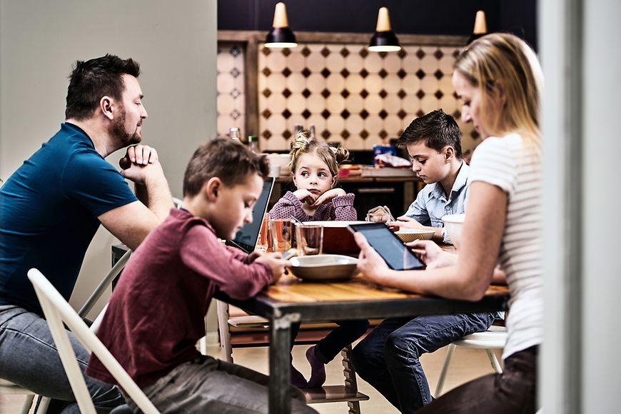 Vater, Mutter und drei Kinder sitzen am Esstisch, Bis auf ein Kind gucken alle anderen auf ihr Handy, Tablet oder den Laptop