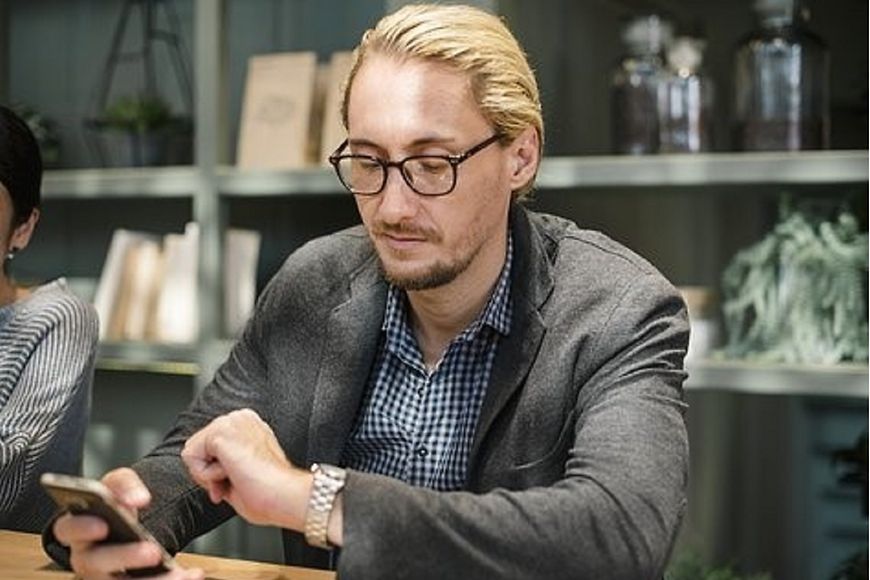 Mann am Tisch mit Handy in der Hand - hinter ihm ein Regal