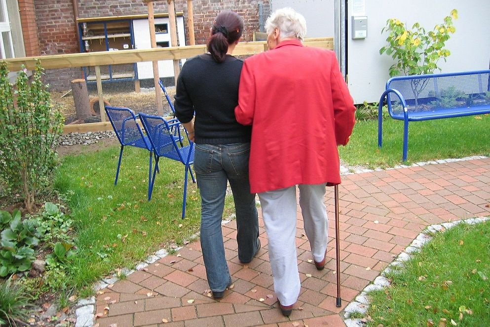 Jüngere und ältere Frau mit Stock gehen einen gepflasterten Weg entlang