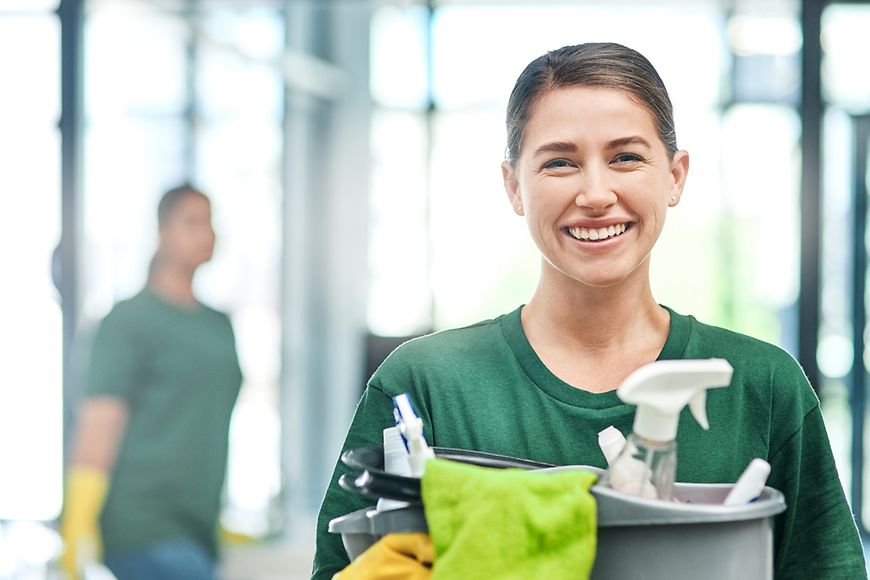 Frau, grünes T-Shirt, lächelt, hat einen Eimer mit Putzmitteln in der Hand, im Hintergrund verschwommene Person