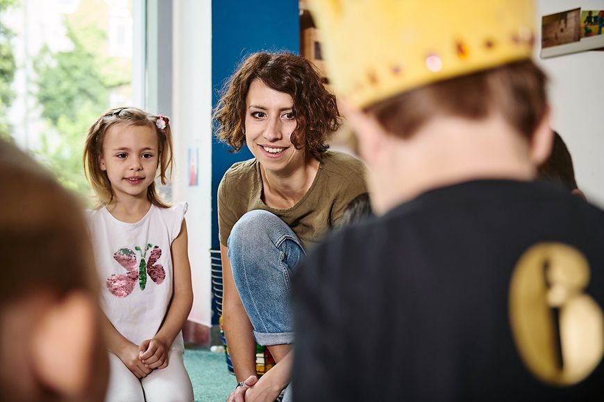 Kitaalltag - Mitarbeiterin, Mädchen und Junge mit Kopfbedeckung sitzen auf dem Boden