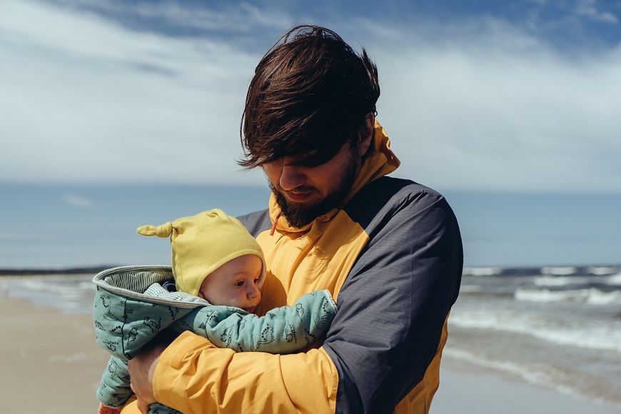 Vater mit Baby auf dem Arm, im Hintergrund sind Strand und Meer zusehen