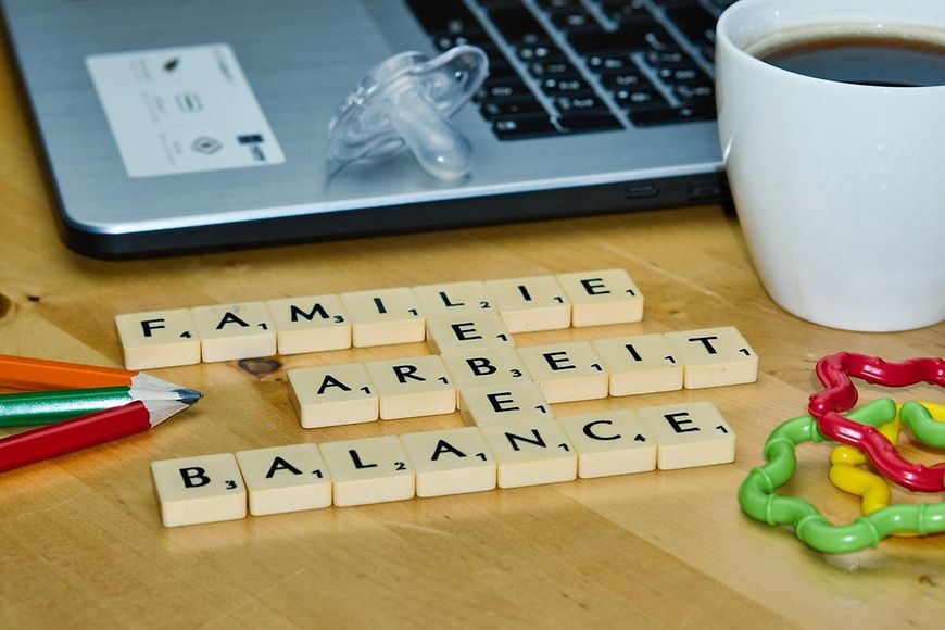Schreibtisch mit Laptop, Kaffeetasse, Babyspielzeug, Stifte und Scrabble-Steinen: Familie, Arbeit, Balance, Leben