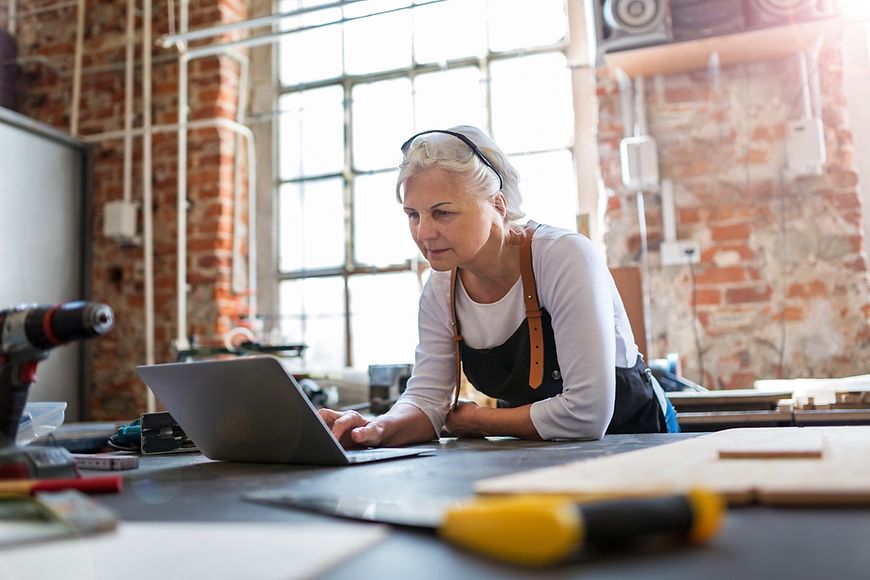 Frau (älter) am Werk- bzw. Zeichentisch guckt auf einen aufgeklappten Laptop