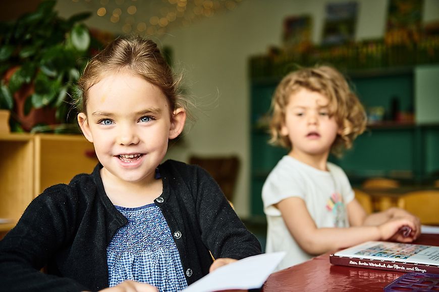 Zwei Kinder sitzen am Tisch, das vordere guckt direkt in die Kamera