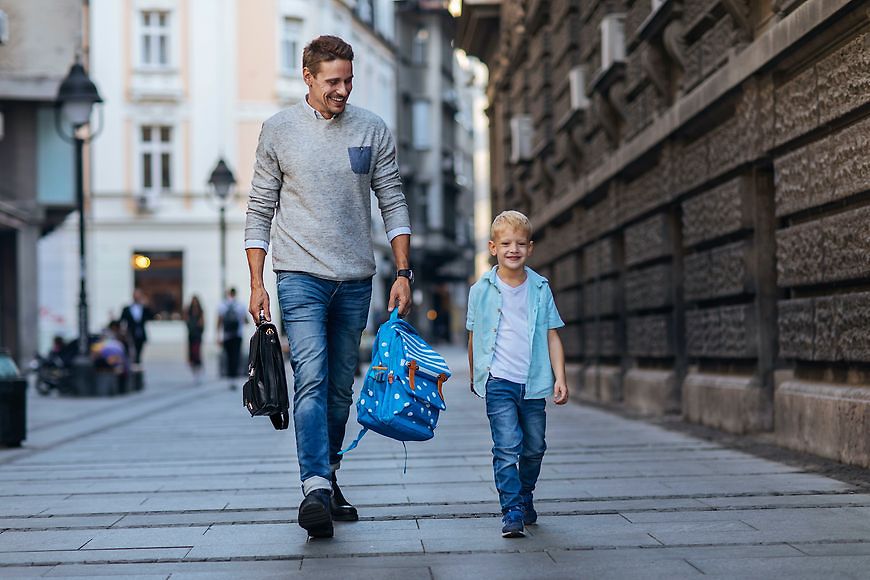 Mann geht mit einem Kind die Straße entlang. Der Mann trägt eine Aktentasche und einen Kinderrucksack. Beide Personen lächeln.