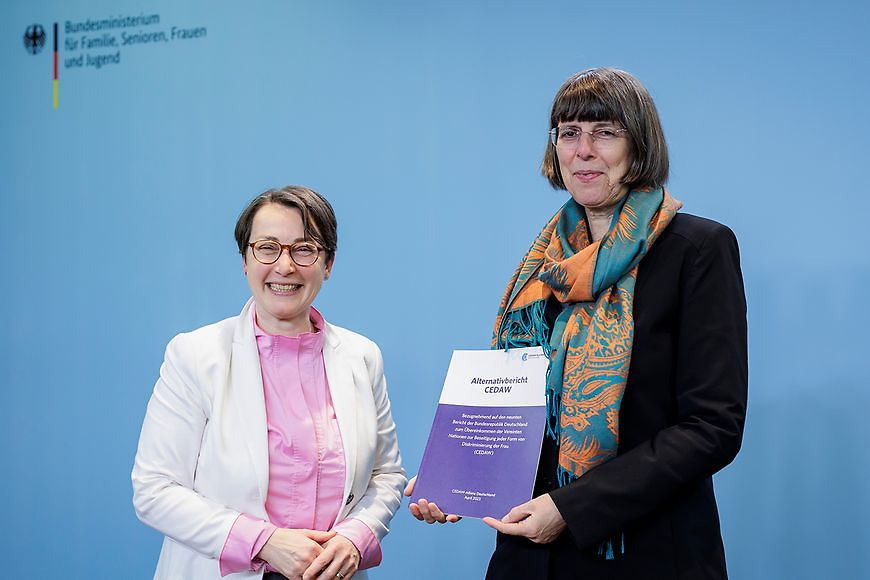 Margit Gottstein hält den Alternativbericht in der Hand, neben ihr steht eine weitere Person und lächelt
