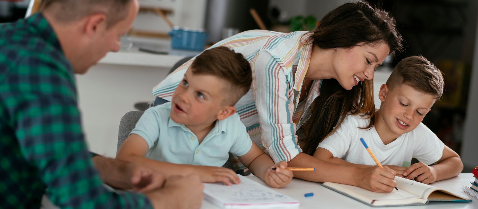 Ein Mann und eine Fraue erklären zwei Kindern etwas, die am Tisch sitzen und Block und Stift in der Hand haben