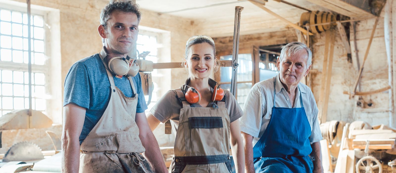 Zwei Männer und eine Frau unterschiedlichen Alters in Arbeitskleidung in einer Werkstatt