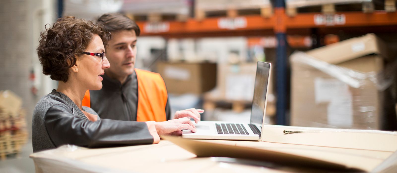Eine Frau und in Mann stehen in einem Lager und gucken gemeinsam auf einen Laptop, der Mann trägt eine orangefarbene Weste