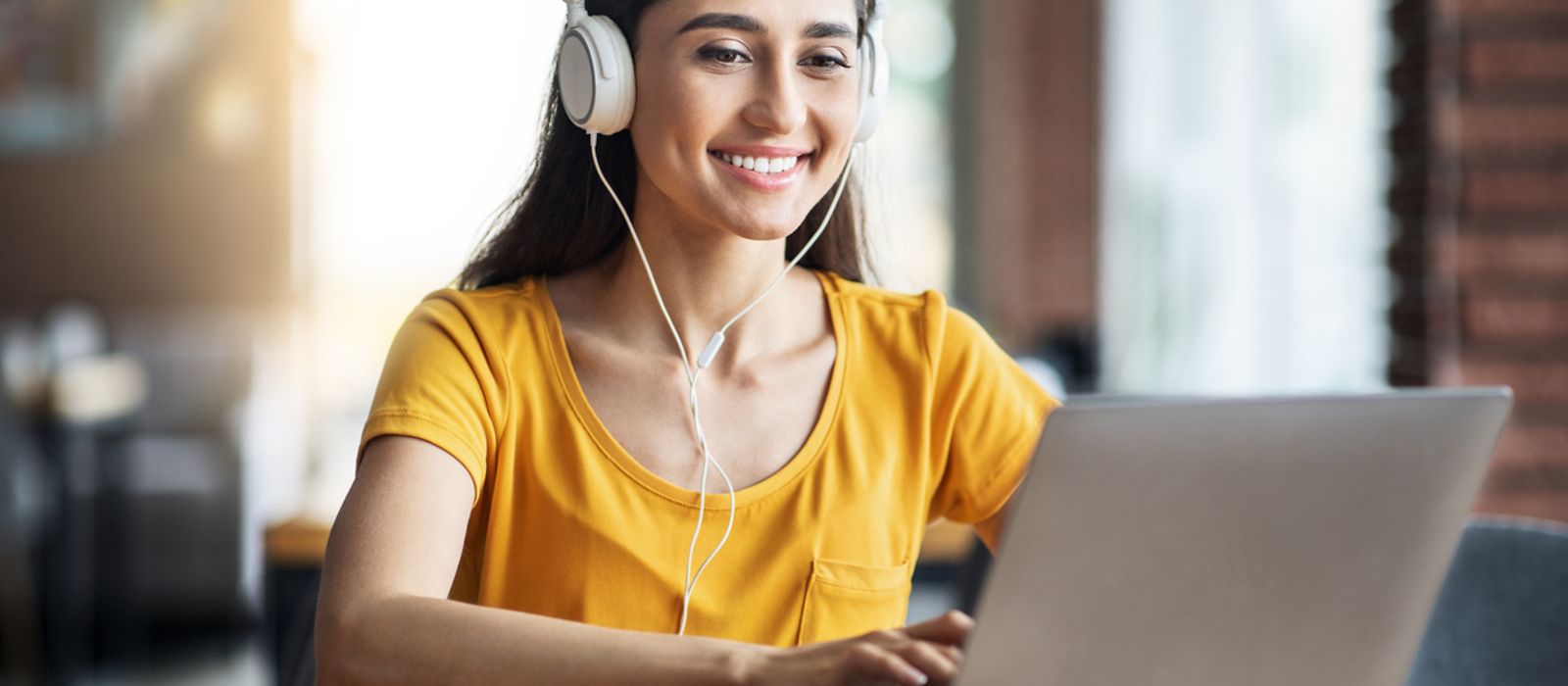 Junge Frau mit Kopfhörern sitzt lächelnd vor einem aufgeklapptem Laptop