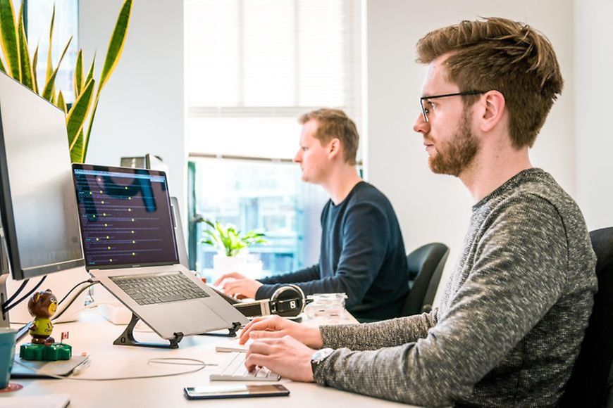 Zwei Männer sitzen am Computer und blicken beide auf ihre jeweiligen Bildschirme