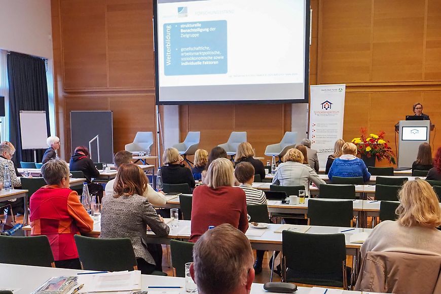 Sitzendes Publikum einer Podiumsdisskussion von hinten, die Bühne mit vier Stühlen ist noch leer