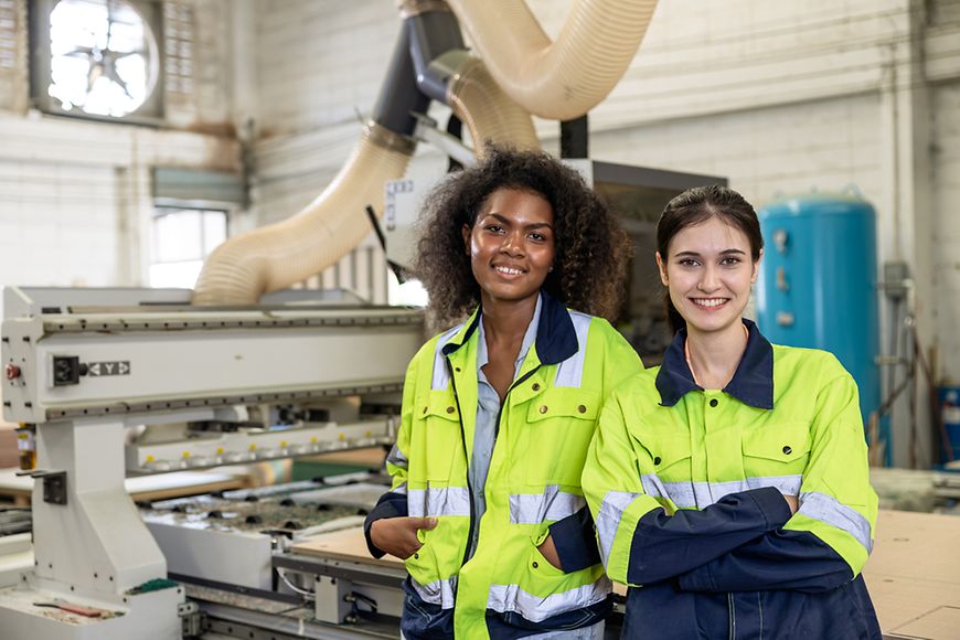 Zwei Frauen in Arbeitskleidung stehen in einer Industriehalle. 