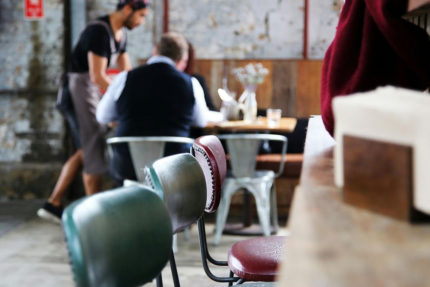 Innenraum eines Restaurants, im Vordergrund leere Stühle, im Hintergrund wird ein Tische bedient